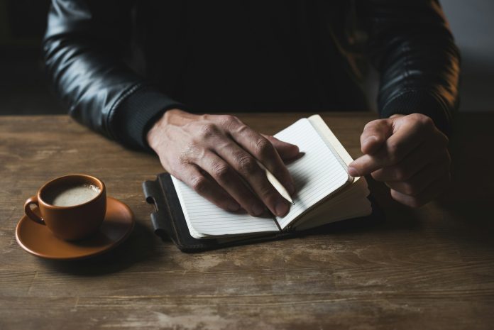 Close-up view of person sitting at wooden table with diary and cup of coffee